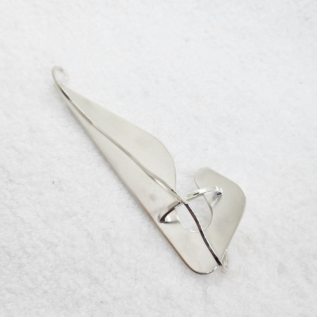 A polished silver pendant with an abstract, organic leaf-like shape and a central loop, displayed on a textured white background.