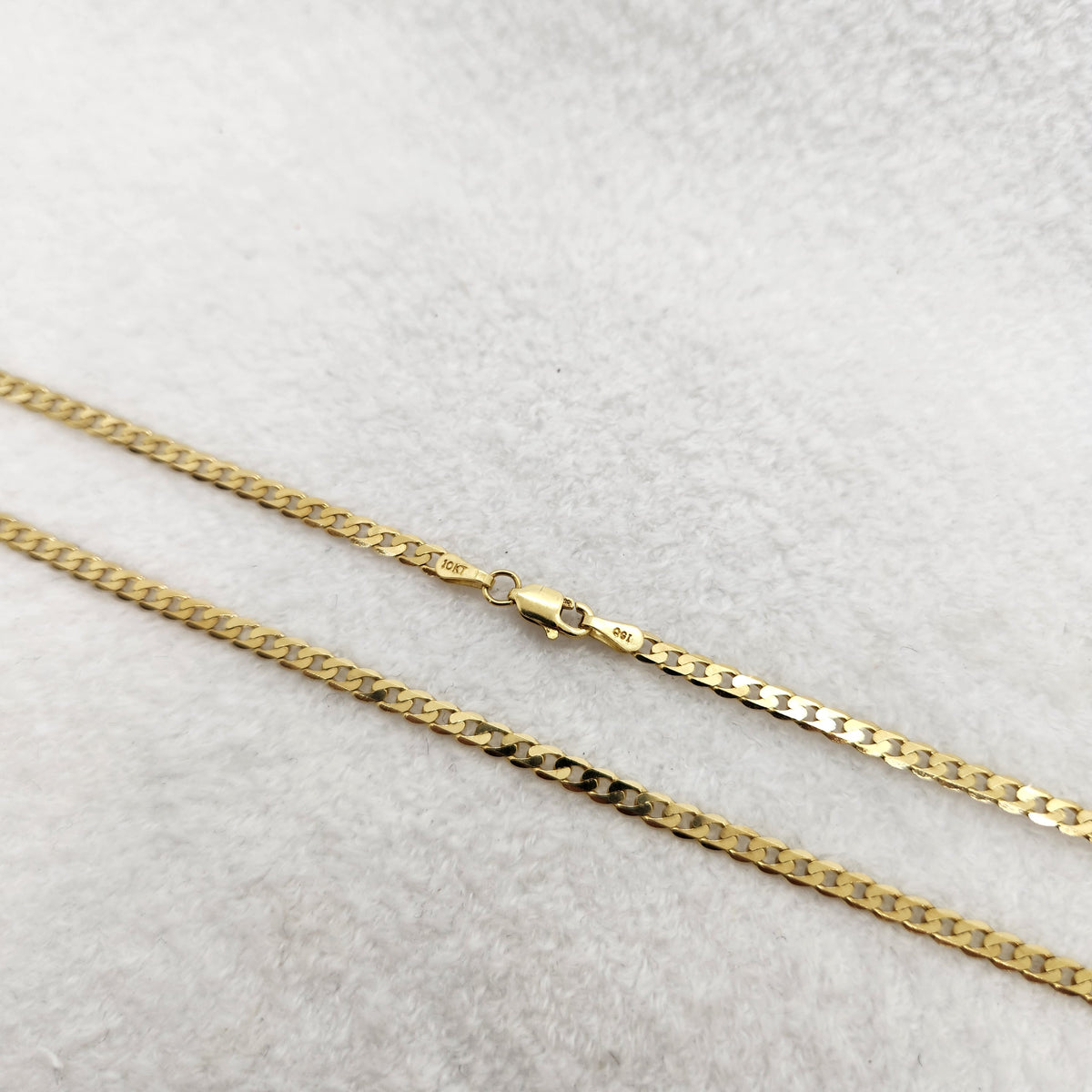 A close-up of a yellow gold chain necklace with a curb link pattern and a lobster clasp, laid diagonally on a textured white fabric background.