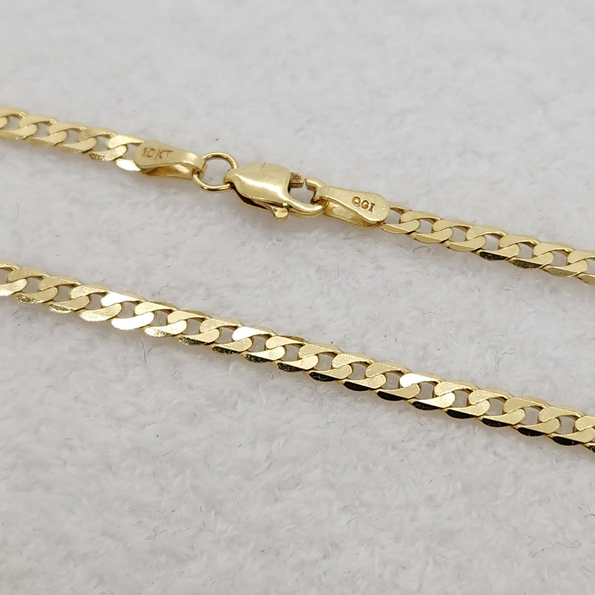 A close-up of a yellow gold chain necklace with a lobster clasp, showing the engraved '10KT' and '925' marks, resting on a textured white surface.