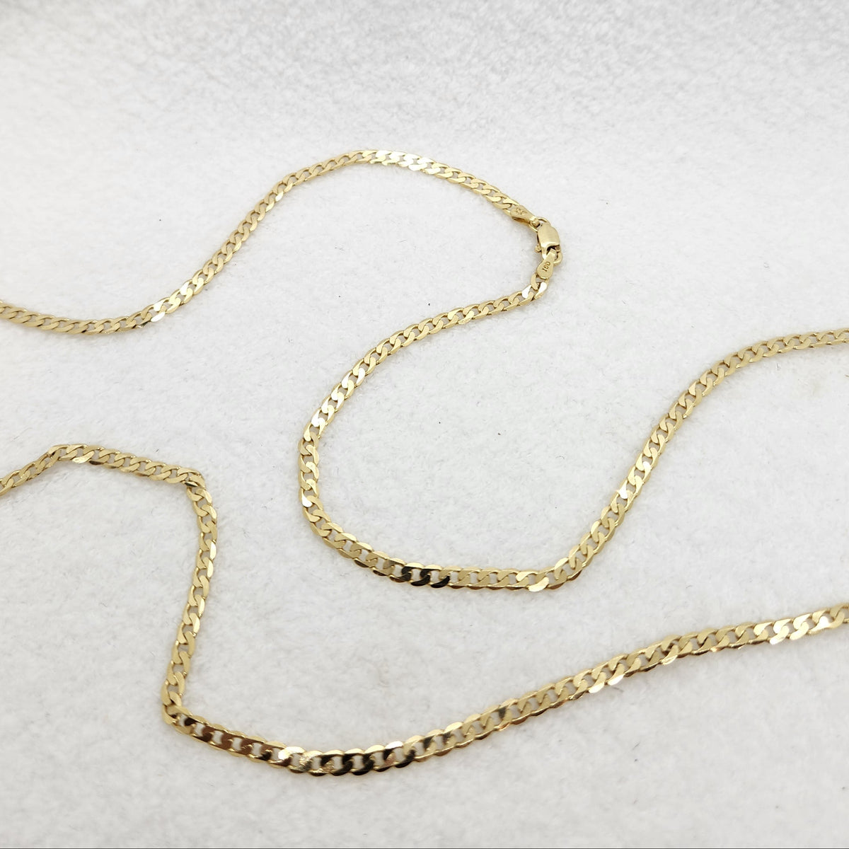 A gold chain necklace with a curb link pattern, laid out in a loose loop on a textured white fabric background.