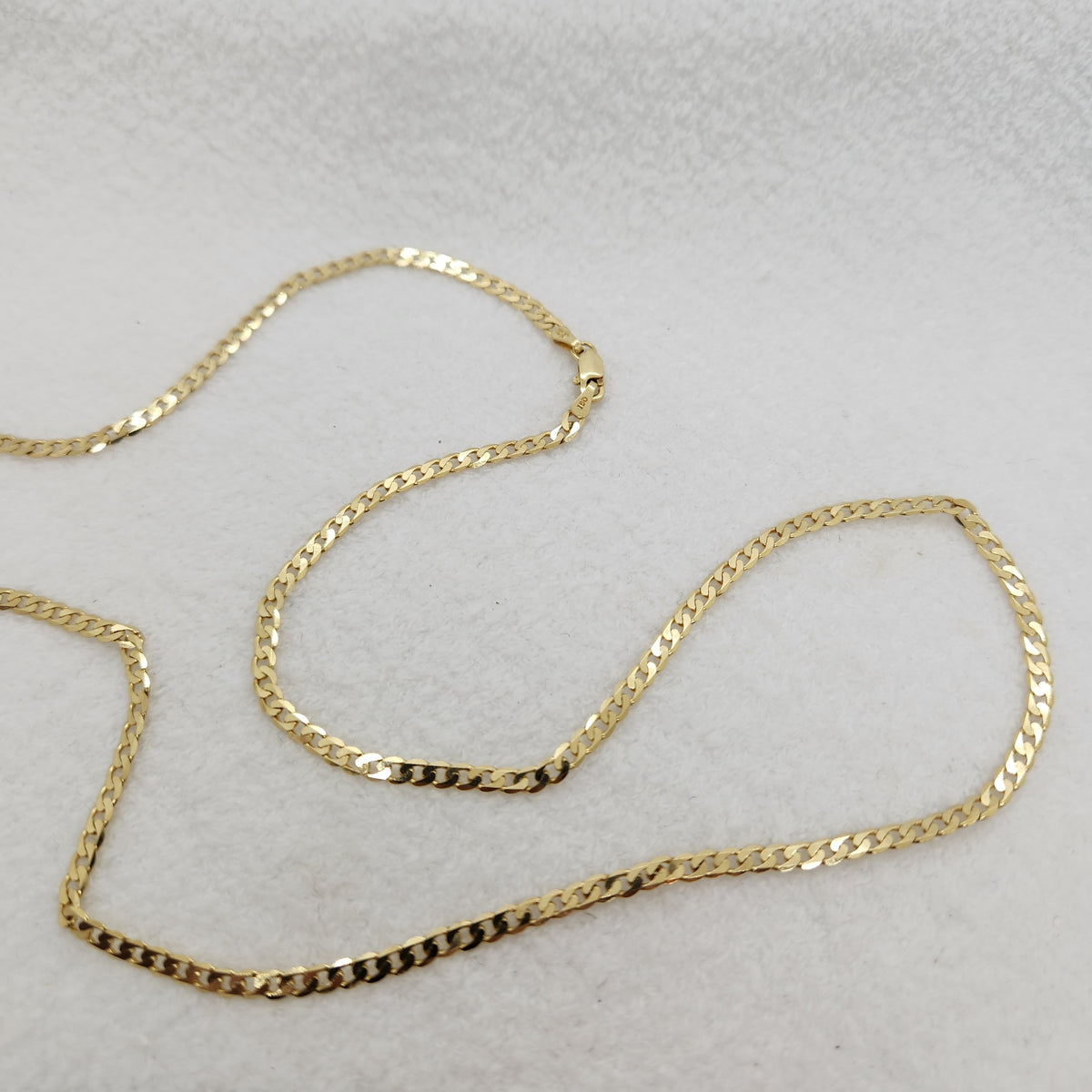 A gold-colored chain necklace, a curb link style, draped in an S-shape on a textured white fabric background.