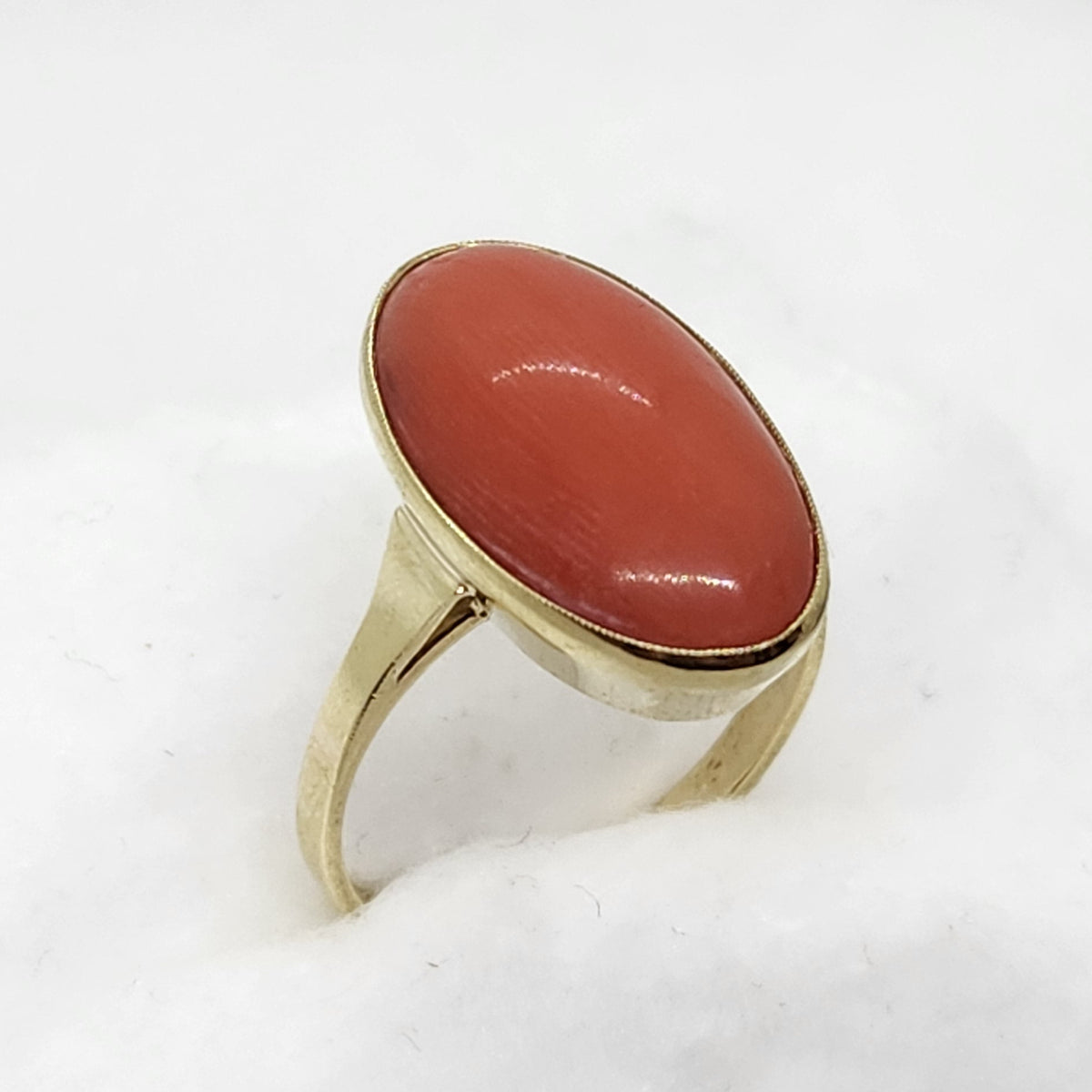 A gold ring with an oval red-orange coral gemstone, displayed on a white textured stand against a white background.