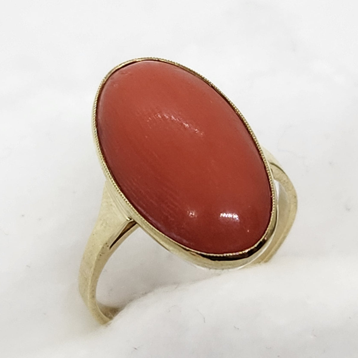 A gold ring with a large, smooth, oval-shaped reddish-orange coral cabochon gemstone, displayed on a white textured stand.