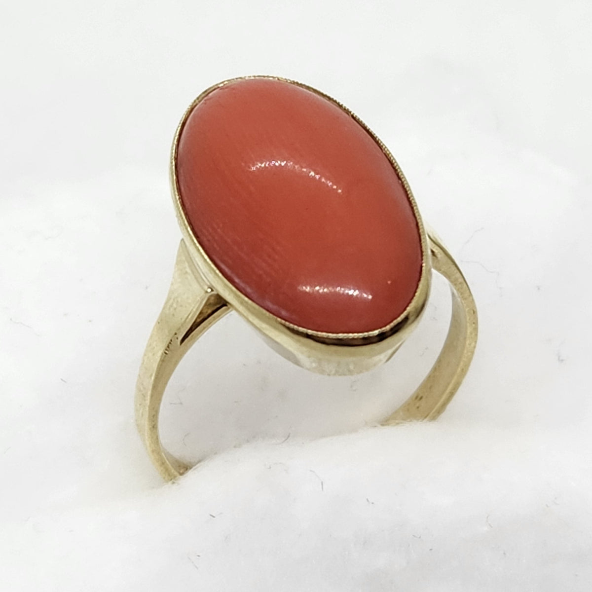 A gold ring with a smooth, polished oval coral cabochon gemstone in a warm reddish-orange hue, displayed on a white textured stand against a white background.