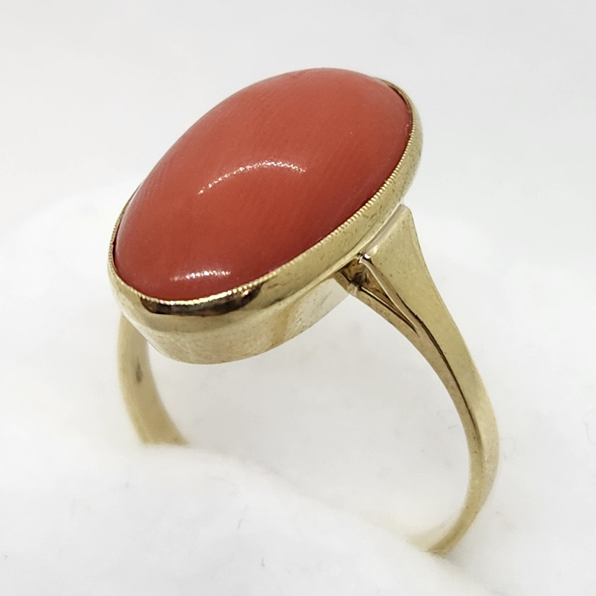 A gold ring featuring a smooth, oval-shaped coral cabochon stone, displayed on a soft white surface against a light gray background.
