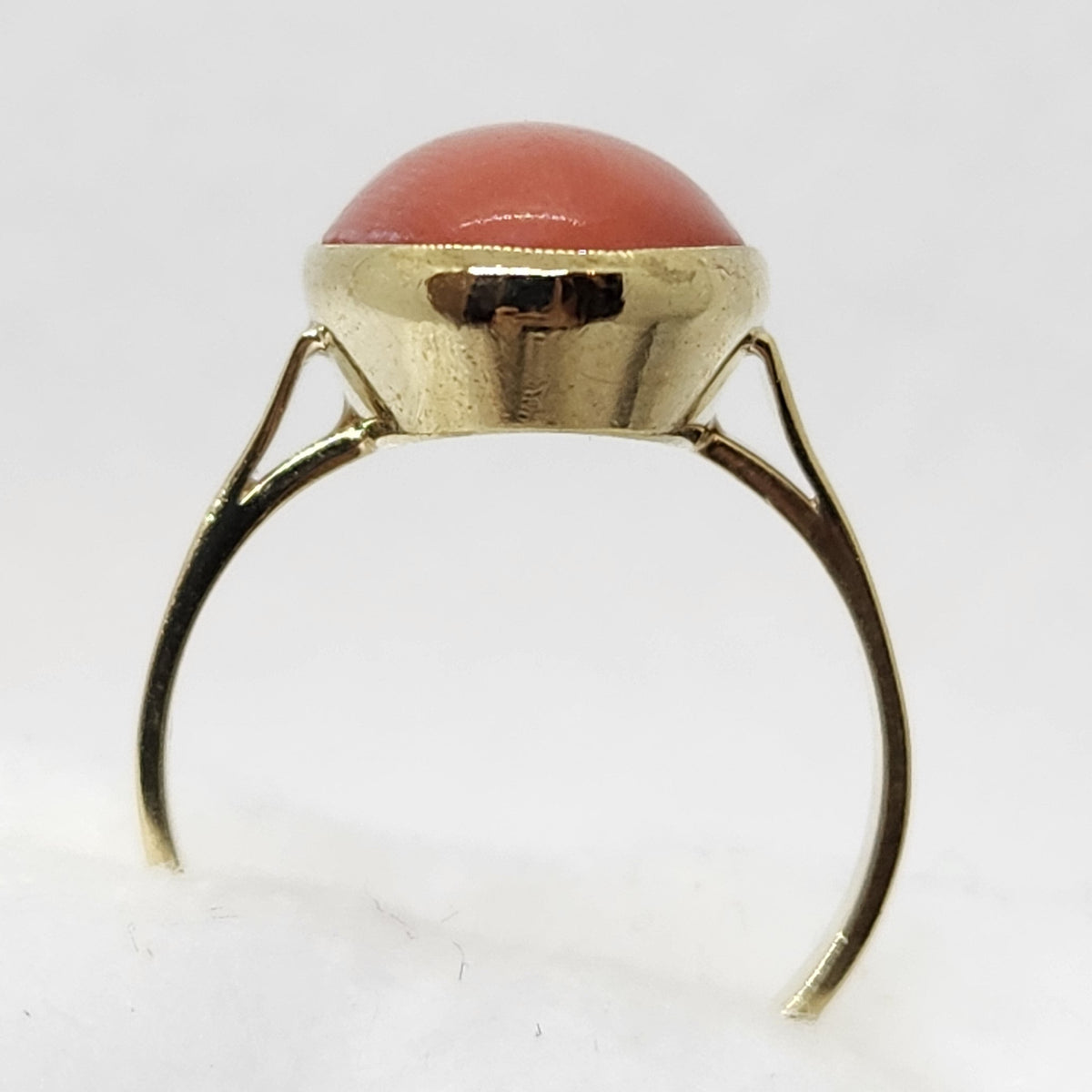 A vintage-style ring featuring a smooth, dome-shaped reddish-orange coral cabochon stone set in a polished gold-tone band, displayed on a soft white textured surface.