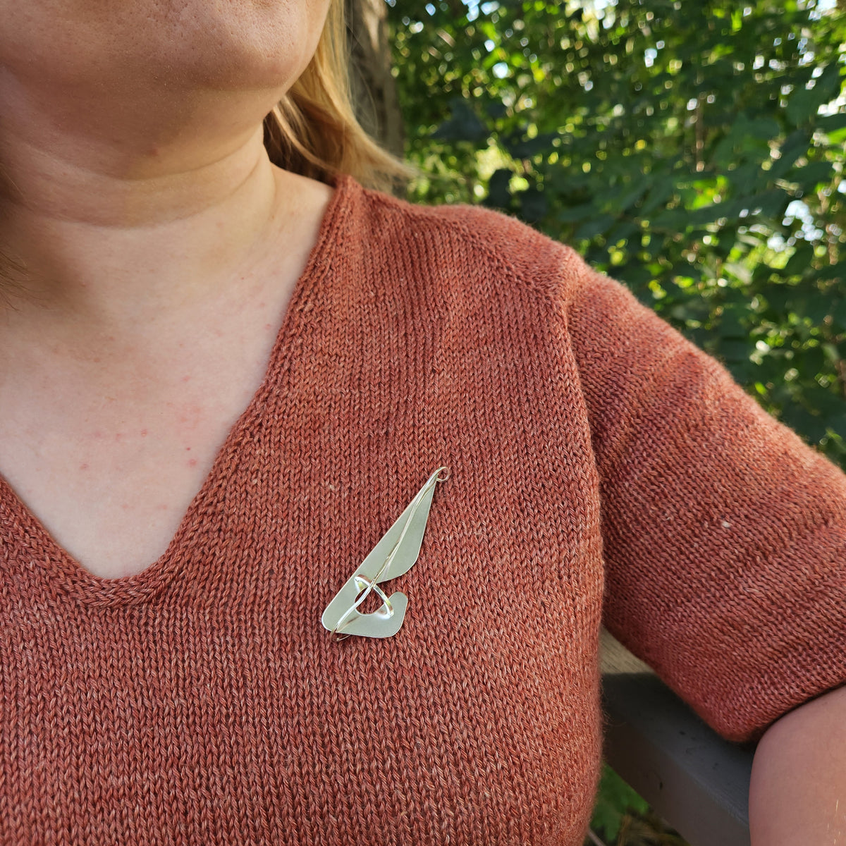 A close-up lifestyle shot of a woman wearing a silver brooch on her chest, wearing a rust-colored knit sweater with a blurred green outdoor background.