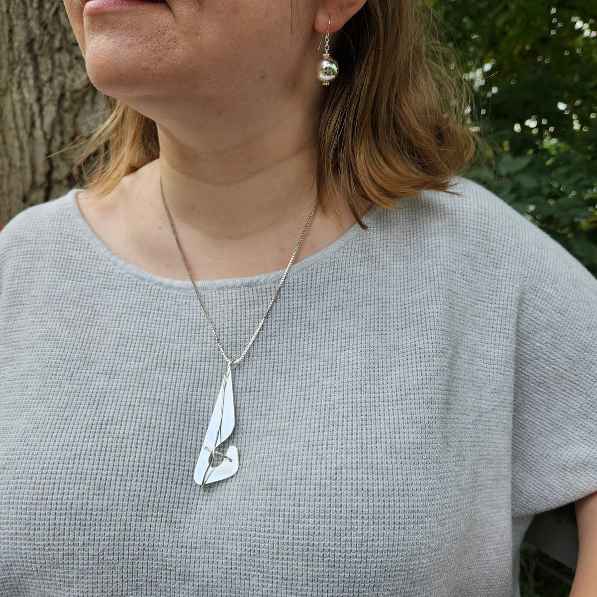 A close-up of a woman wearing a silver pendant necklace with a stylized leaf or feather design, paired with a matching silver drop earring, set against a background of a tree trunk and green foliage.