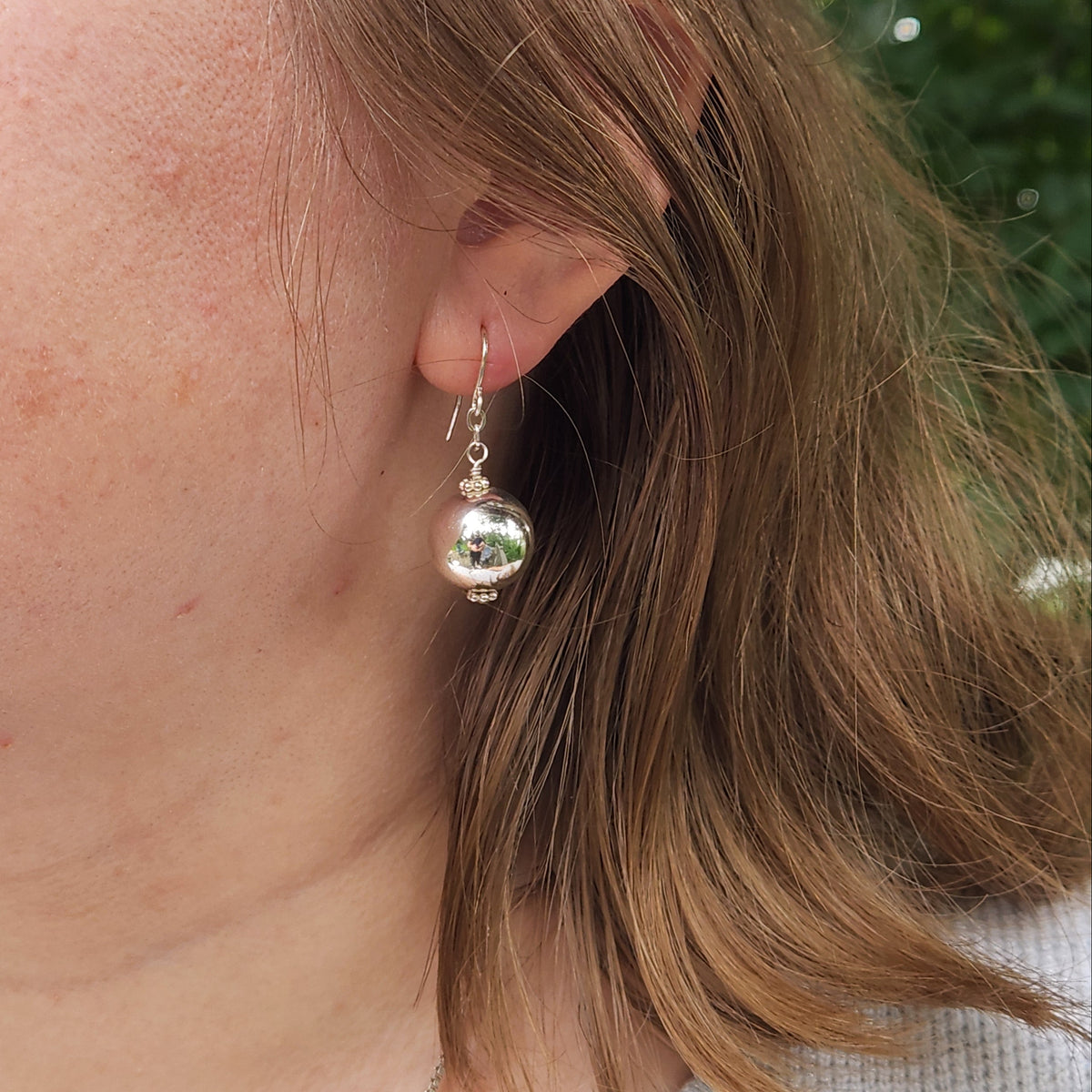 A close-up of a person's ear wearing a silver dangle earring with a small reflective sphere, against a blurred outdoor background.