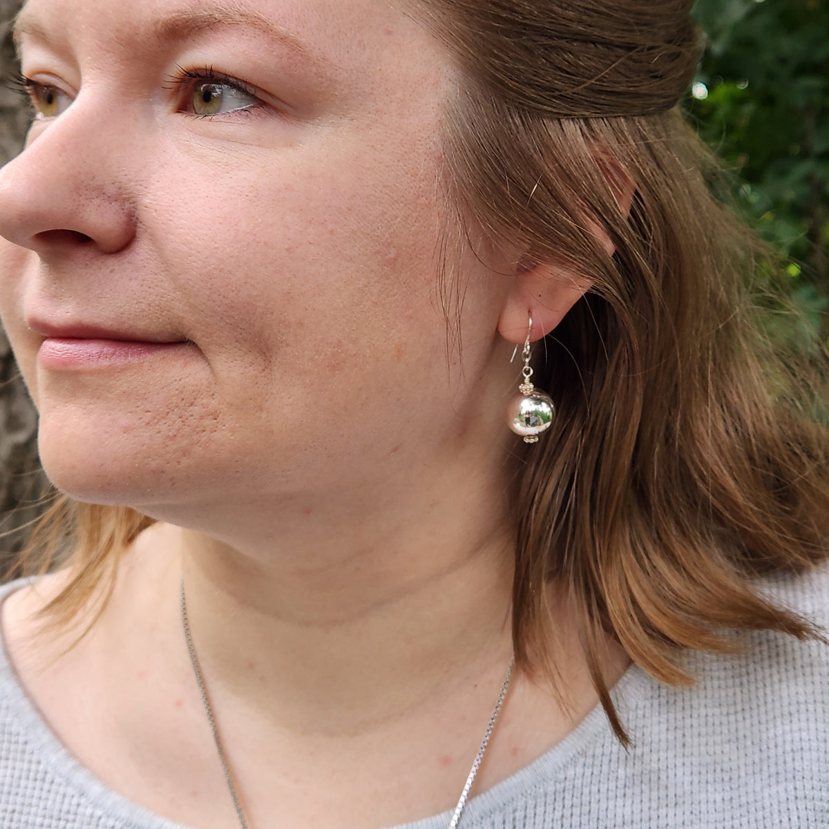 A close-up side profile of a woman wearing a silver dangle earring with a clear faceted bead, set against an outdoor background with soft green foliage.