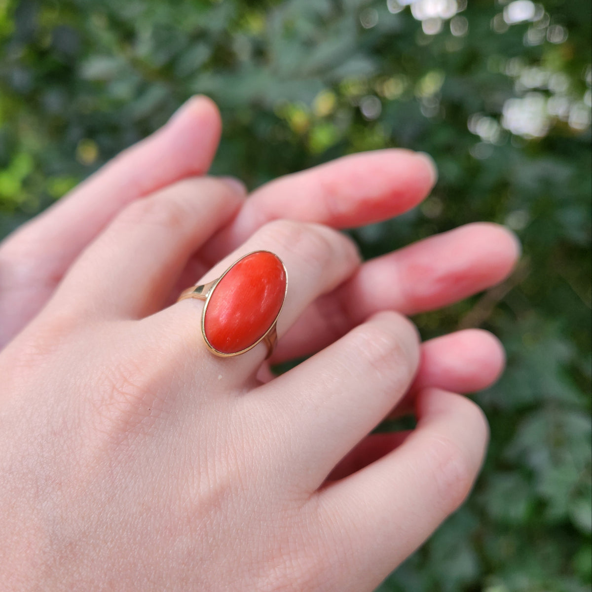 A gold ring with a large oval orange-red cabochon coral gemstone worn on a person's finger, photographed outdoors against a blurred green foliage background.