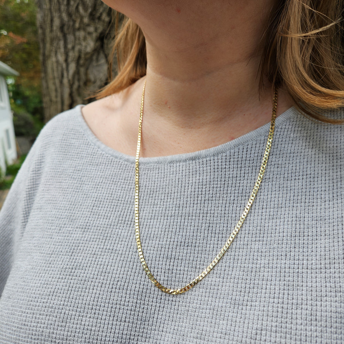 A close-up of a person wearing a delicate gold chain necklace over a light gray ribbed knit top, with a blurred outdoor background.
