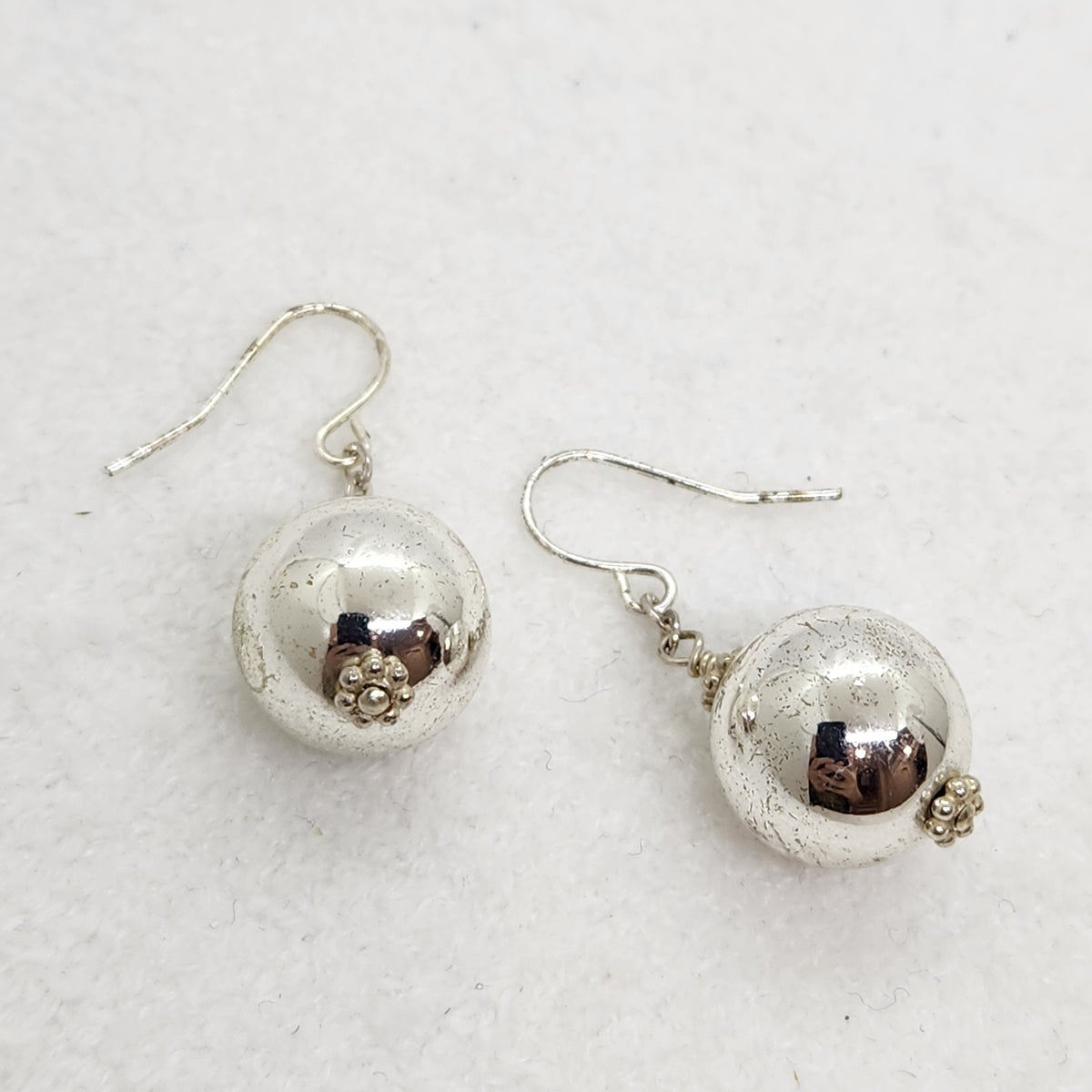A pair of silver dangle earrings with textured spherical beads and small floral accents, displayed on a light textured white background.