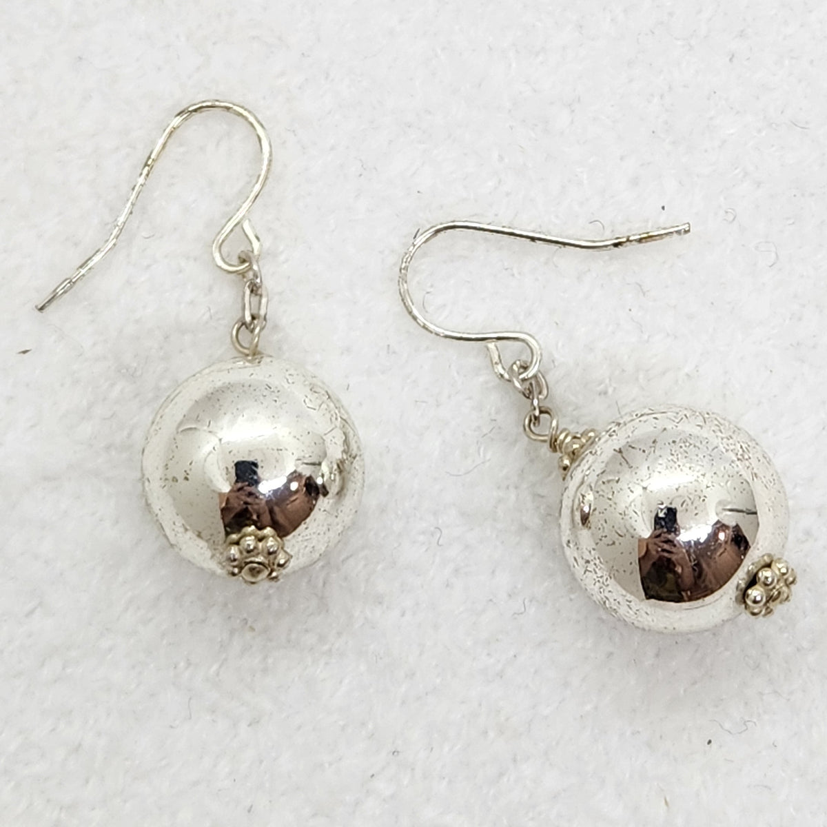 A pair of silver dangle earrings with textured, hammered spherical beads and small floral accents, displayed on a textured white background.