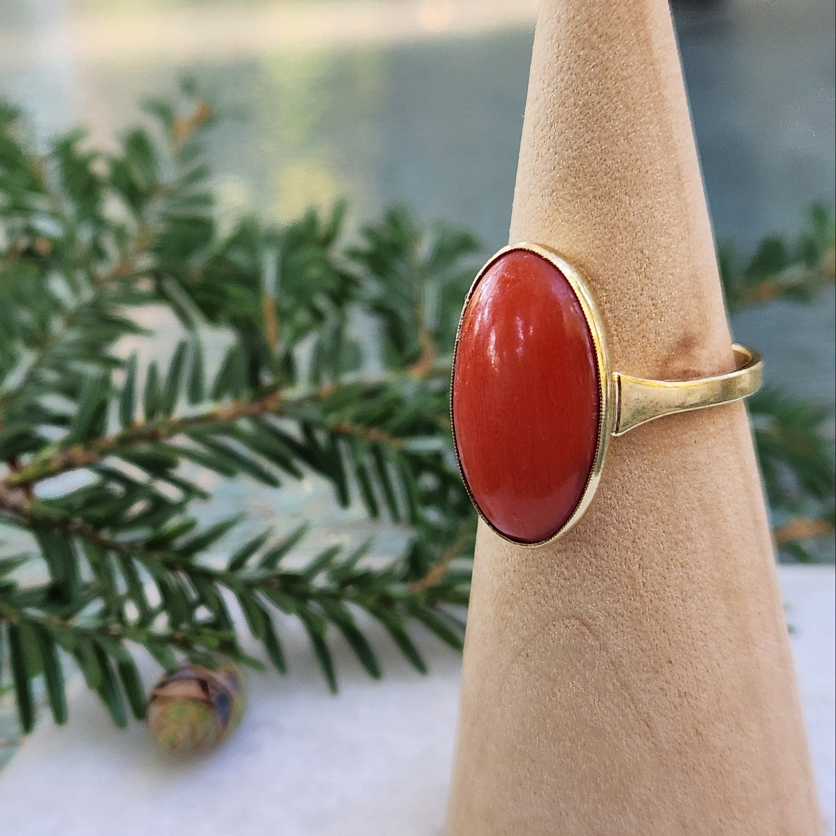 A gold ring with a large oval red coral cabochon gemstone displayed on a cone stand, with green pine branches in the background.