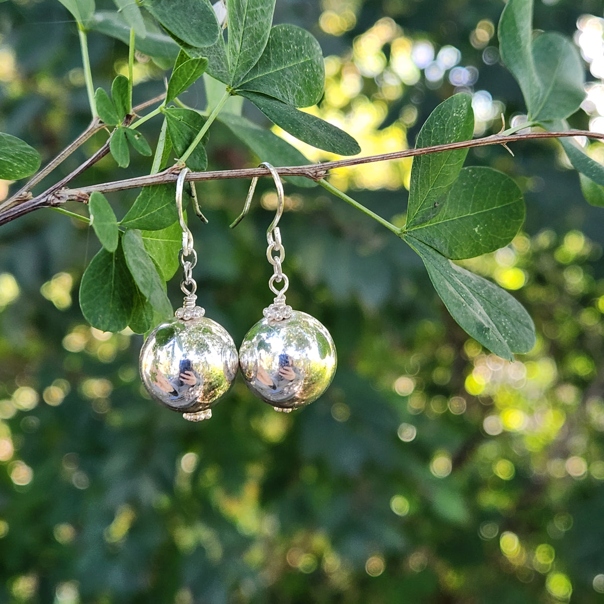 A pair of silver dangle earrings with reflective spherical beads hanging from a tree branch with green leaves, set against a blurred green outdoor background.