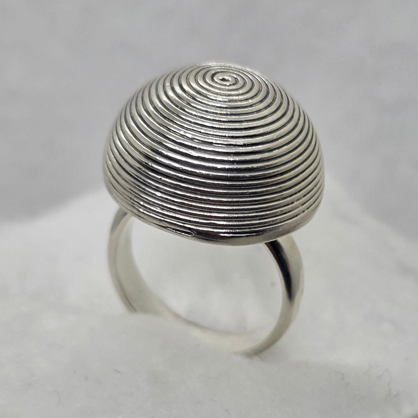 A silver ring with a large, round, spiral-patterned top, displayed on a white textured stand.