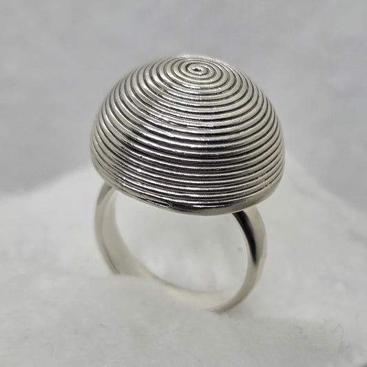 A silver ring with a large, round, spiral-patterned top, displayed on a white textured stand.